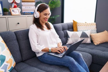 Young beautiful hispanic woman using laptop and headphones sitting on sofa at home