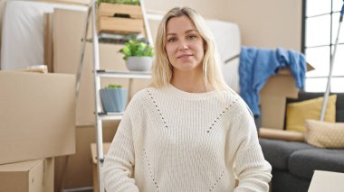 Genç sarışın kadın yeni evinde yerde kendinden emin bir şekilde gülümsüyor.