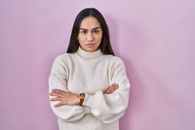 Pembe arka planda duran genç Güney Asyalı kadın şüpheci ve gergin, yüzünde çapraz kollu tasvip etmeyen ifade. negatif kişi. 