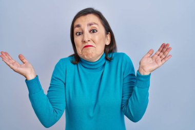 Orta yaşlı İspanyol kadın soyutlanmış arka planda duruyor. Kolları ve elleri havada, kafası karışmış bir ifade. şüphe kavramı. 