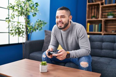 Young latin man playing video game sitting on sofa at home