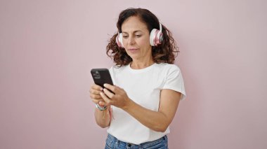 Orta yaşlı İspanyol kadın, pembe arka planda kulaklık takarak akıllı telefon kullanıyor.