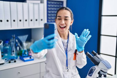 Genç Güney Asyalı bir kadın akıllı telefonuyla laboratuvarda çalışıyor. Pozitif ve mutlu görünüyor. Kendine güvenen bir gülümsemeyle dişlerini gösteriyor. 