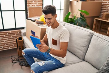 Young hispanic man using touchpad and credit card sitting on sofa at new home