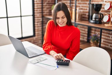 Young beautiful hispanic woman using laptop and calculator sitting on table at home