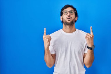 Mavi arka planda duran yakışıklı Latin adam şaşırmış ve şaşırmış bir şekilde yukarı bakıyor ve parmaklarıyla işaret ediyor ve kollarını kaldırıyor.. 