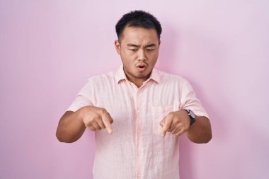 Pembe arka planda duran Çinli genç adam parmaklarını göstererek reklamını yapıyor, şaşırmış yüzünü ve ağzını açıyor. 