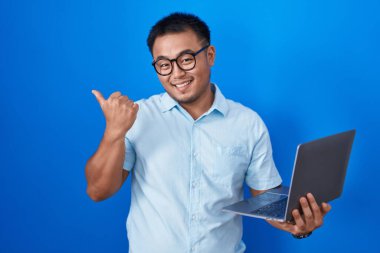 Çinli genç adam bilgisayarla arka tarafı işaret ediyor. Elleri havada, kendinden emin bir şekilde gülümsüyor. 