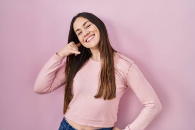 Young brunette woman standing over pink background smiling doing phone gesture with hand and fingers like talking on the telephone. communicating concepts. 