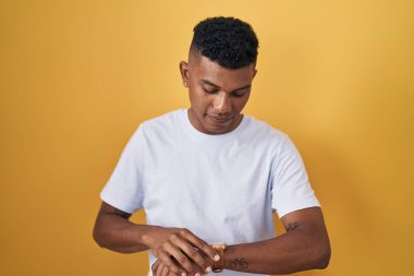 Young hispanic man standing over yellow background checking the time on wrist watch, relaxed and confident 