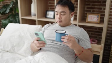 Genç Çinli adam akıllı telefon kullanıyor. Yatak odasında kahve içiyor.