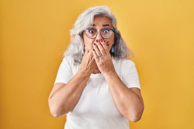 Middle age woman with grey hair standing over yellow background shocked covering mouth with hands for mistake. secret concept. 