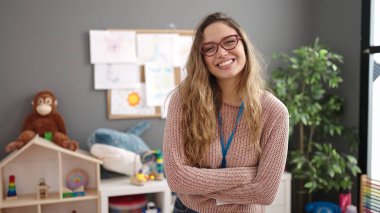 Young beautiful hispanic woman preschool teacher smiling confident standing with arms crossed gesture at kindergarten