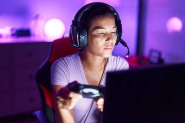 Young beautiful hispanic woman streamer playing video game using joystick at gaming room