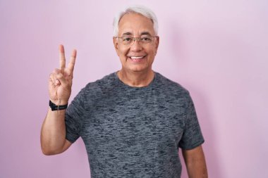 Pembe arka planda duran gri saçlı orta yaşlı bir adam iki numaralı parmaklarıyla işaret ederken kendinden emin ve mutlu gülümsüyor.. 