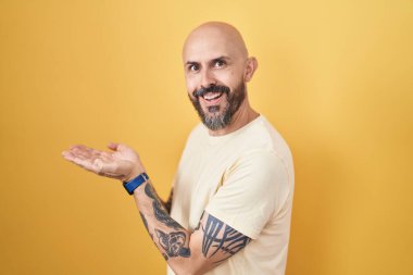 Sarı arka planda dikilen İspanyol dövmeli adam avuçları açık, fotokopi alanını gösteren, gülümseyerek mutlu bir gülümseme sunan. 