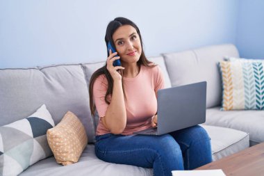 Genç, güzel, İspanyol bir kadın akıllı telefonuyla evde dizüstü bilgisayar kullanıyor.