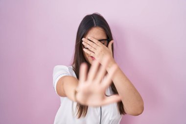 Genç esmer kadın pembe arka planda duran gözlüklü, elleriyle gözlerini kapayan, üzgün ve korku ifadeli hareketlerini durduran. utangaç ve olumsuz kavram. 