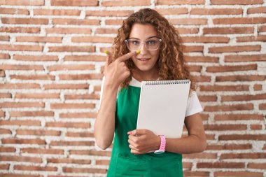 Genç, beyaz bir kadın, elinde sanat defteri tutarken, parmakları burnuna ve yüzüne dönük, neşeli bir şekilde gülümsüyor. güzellik konsepti 