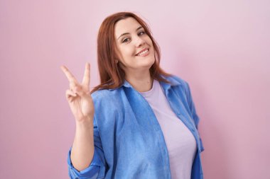 Pembe arka planda duran, gülümseyen, parmaklarını zafer işareti yapan Latin kökenli genç bir kadın. İki numara.. 