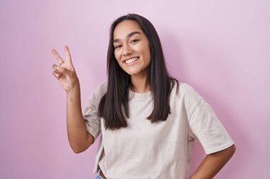 Young hispanic woman standing over pink background smiling looking to the camera showing fingers doing victory sign. number two. 