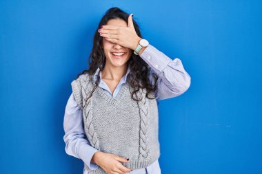 Genç esmer kadın mavi arka planda duruyor. Gülümsüyor ve gülümsüyor. Gözleri şaşkınlıkla örtüyor. kör kavram. 