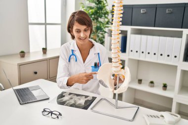 Young beautiful hispanic woman doctor pointing with finger to anatomical model of vertebral column at clinic