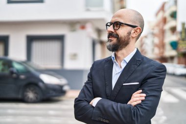 Kel genç bir iş adamı caddede kolları çapraz hareketlerle duruyor.