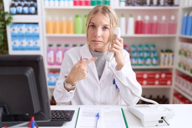 Eczanede çalışan beyaz bir kadın telefonda konuşuyor. Endişeli ve endişeli bir ifadeyle işaret ediyor. 