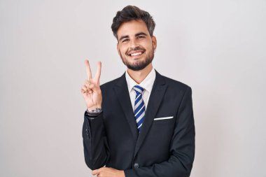İş elbisesi ve kravat takan, gülümseyen, kameraya göz kırpan, parmaklarıyla zafer işareti yapan İspanyol dövmeli genç adam. İki numara.. 