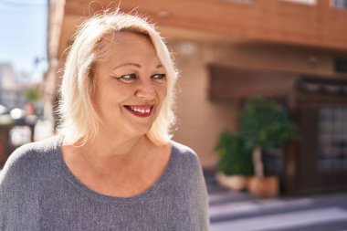Orta yaşlı sarışın kadın sokakta kendinden emin bir şekilde gülümsüyor.