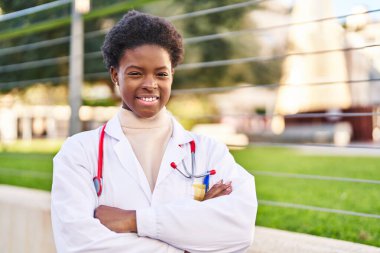 Doktor üniforması giyen Afrikalı Amerikalı kadın caddede kollarını kavuşturmuş duruyor.