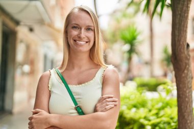 Genç sarışın kadın gülümsüyor. Kollarını kavuşturup caddede duruyor.