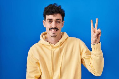 Mavi arka planda dikilen İspanyol adam kameraya gülümseyip parmaklarıyla zafer işareti yapıyor. İki numara.. 