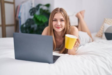 Laptop kullanan, yatakta kahve içen beyaz bir kadın.