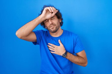 İspanyol genç adam mavi arka planda duruyor. Hastalık, ateş, grip ve virüs için alnına dokunuyor. 