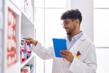 Young arab man pharmacist using touchpad holding medicine bottle at pharmacy