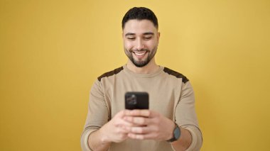 Genç Arap adam kendinden emin bir şekilde sarı arka plan üzerinde akıllı telefon kullanıyor.