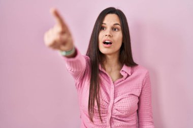 Young hispanic woman standing over pink background pointing with finger surprised ahead, open mouth amazed expression, something on the front 