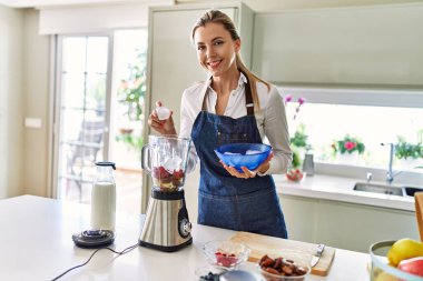 Genç sarışın kadın mutfaktaki blendere buz dökerek gülümsüyor.