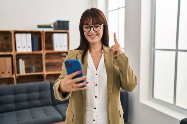 Ofiste çalışan genç esmer kadın akıllı telefonuyla gülümsüyor bir fikir veya soru işareti ile mutlu bir yüzle, bir numara 
