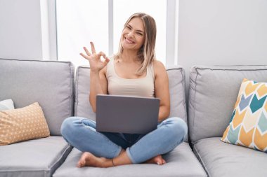 Sarışın, beyaz bir kadın evinde dizüstü bilgisayar kullanıyor. Kanepede oturuyor. Parmaklarıyla işaret veriyor. 