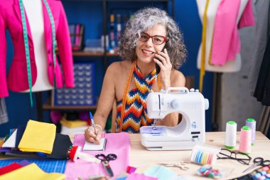 Middle age grey-haired woman tailor talking on smartphone writing on notebook at sewing studio