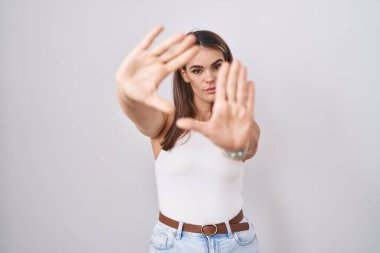 Latin kökenli genç bir kadın beyaz arka planda duruyor. El ve parmakları kullanarak çerçeve yapıyor. Kamera bakış açısı. 