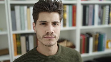 Young hispanic man student sitting on table with relaxed expression at library university