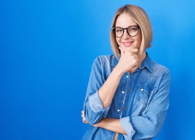 Mavi arka planda duran genç beyaz kadın kameraya kollarını kavuşturup gülümseyerek ve çenesini kaldırarak bakıyor. Olumlu düşünmek. 