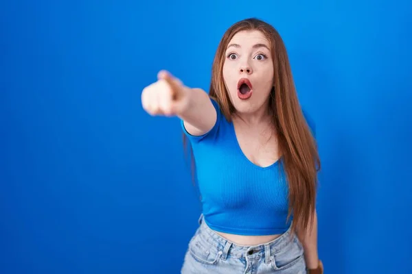 赤頭の女性の青の背景の上に指を前に 口を開けて驚いて表現 何かの前に立っている — ストック写真