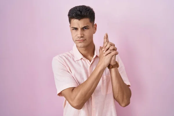 Young hispanic man standing over pink background holding symbolic gun with hand gesture, playing killing shooting weapons, angry face