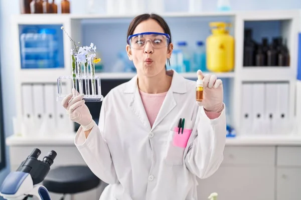 Young brunette woman working at scientist laboratory making fish face with mouth and squinting eyes, crazy and comical.