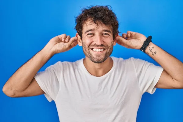 西班牙裔年轻人站在蓝色的背景上 微笑着用手指拉耳朵 做着滑稽的手势 试镜问题 — 图库照片